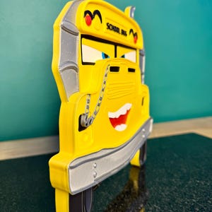 K&ouml;nnte beinhalten: Ein gelbes, schulbusf&ouml;rmiges Dekorationsst&uuml;ck mit einem l&auml;chelnden Gesicht und dem Schriftzug "SCHOOL BUS" an der Seite. Der Bus hat schwarze Reifen, silberne Akzente und eine Kettendetail. Die Augen sind blau und der Mund rot.
