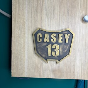 May include: A custom-shaped plaque with a metallic bronze finish featuring the name CASEY and the number 13 in raised, bold lettering. The plaque has a shield-like silhouette with a dark, weathered texture and is mounted on a light wood surface.