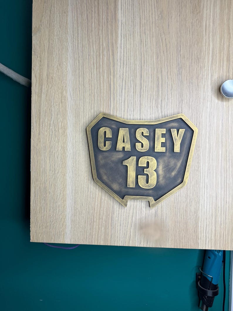 May include: A custom-shaped plaque with a metallic bronze finish featuring the name CASEY and the number 13 in raised, bold lettering. The plaque has a shield-like silhouette with a dark, weathered texture and is mounted on a light wood surface.
