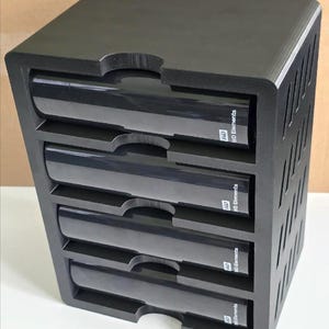 Puede incluir: Una unidad de almacenamiento negra con cinco ranuras horizontales, cada una con un disco duro WD Elements. La unidad tiene forma rectangular con una parte superior y laterales sólidos, y una serie de ranuras verticales en el lado derecho para ventilación.