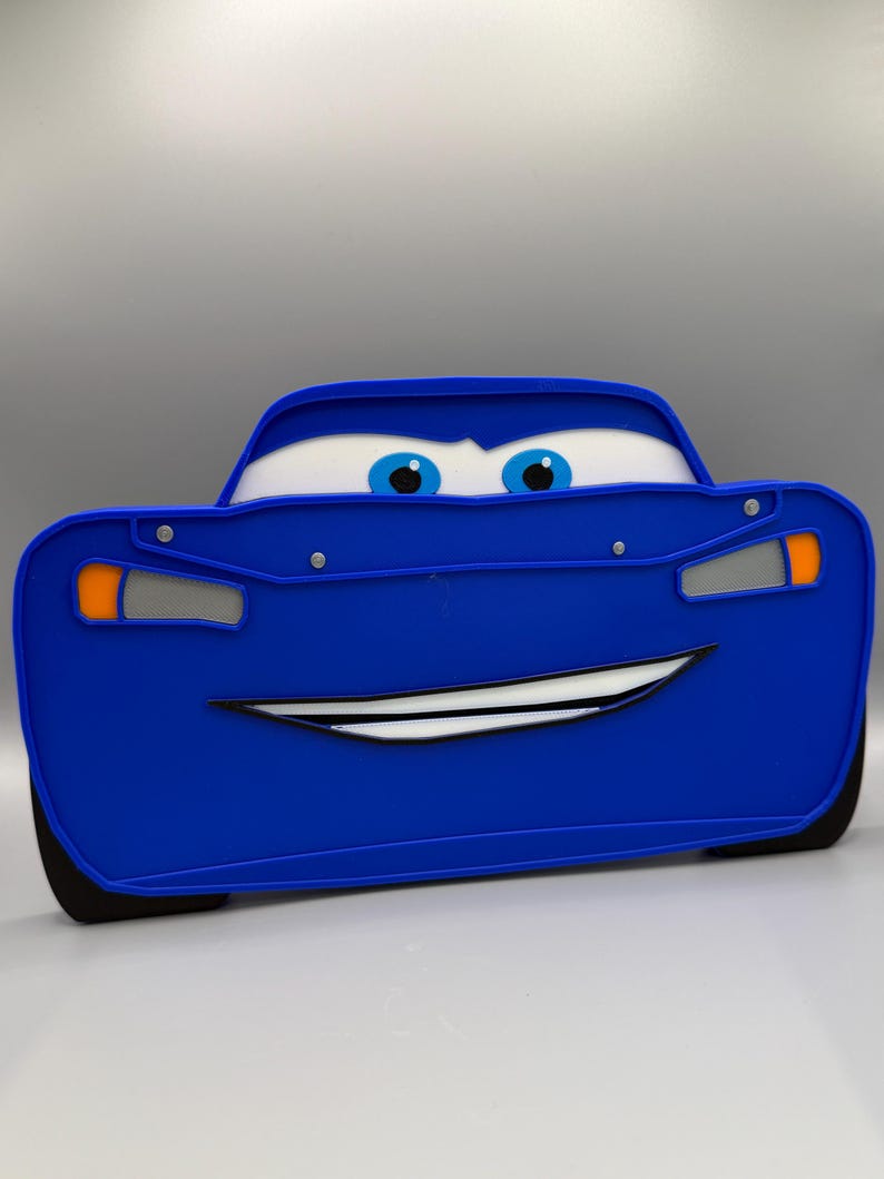 May include: A blue Lightning McQueen-themed toy car with a smiling face, white eyes, and blue irises. The car has black tires and orange and silver accents. The toy is designed to resemble the character from the animated movie "Cars".