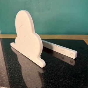 May include: A white, cloud-shaped decorative item with a rectangular support. The cloud has a rounded shape with three curved indentations. The support has three circular holes. The item is sitting on a black, speckled surface.