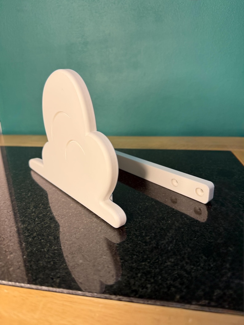 May include: A white, cloud-shaped decorative item with a rectangular support. The cloud has a rounded shape with three curved indentations. The support has three circular holes. The item is sitting on a black, speckled surface.