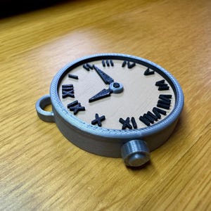 May include: A silver-coloured pocket watch with Roman numeral markings on the face. The watch has a black hour hand and a black minute hand. The watch is on a wooden surface.