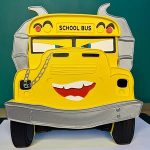 K&ouml;nnte beinhalten: Ein gelber Schulbus mit einem l&auml;chelnden Gesicht, schwarzen Reifen und einer silbernen Sto&szlig;stange. Der Bus tr&auml;gt die Aufschrift "SCHOOL BUS" in schwarzer Schrift. Der Bus hat eine Kette an der Vorderseite und einen rot-wei&szlig;en Mund.