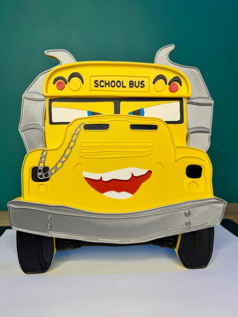 K&ouml;nnte beinhalten: Ein gelber Schulbus mit einem l&auml;chelnden Gesicht, schwarzen Reifen und einer silbernen Sto&szlig;stange. Der Bus tr&auml;gt die Aufschrift "SCHOOL BUS" in schwarzer Schrift. Der Bus hat eine Kette an der Vorderseite und einen rot-wei&szlig;en Mund.