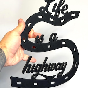 May include: A black wall sign shaped like a winding road with dashed white-style markings, featuring the cut-out text Life is a highway in a cursive script font. The object is constructed from a flat, solid material with a matte finish.