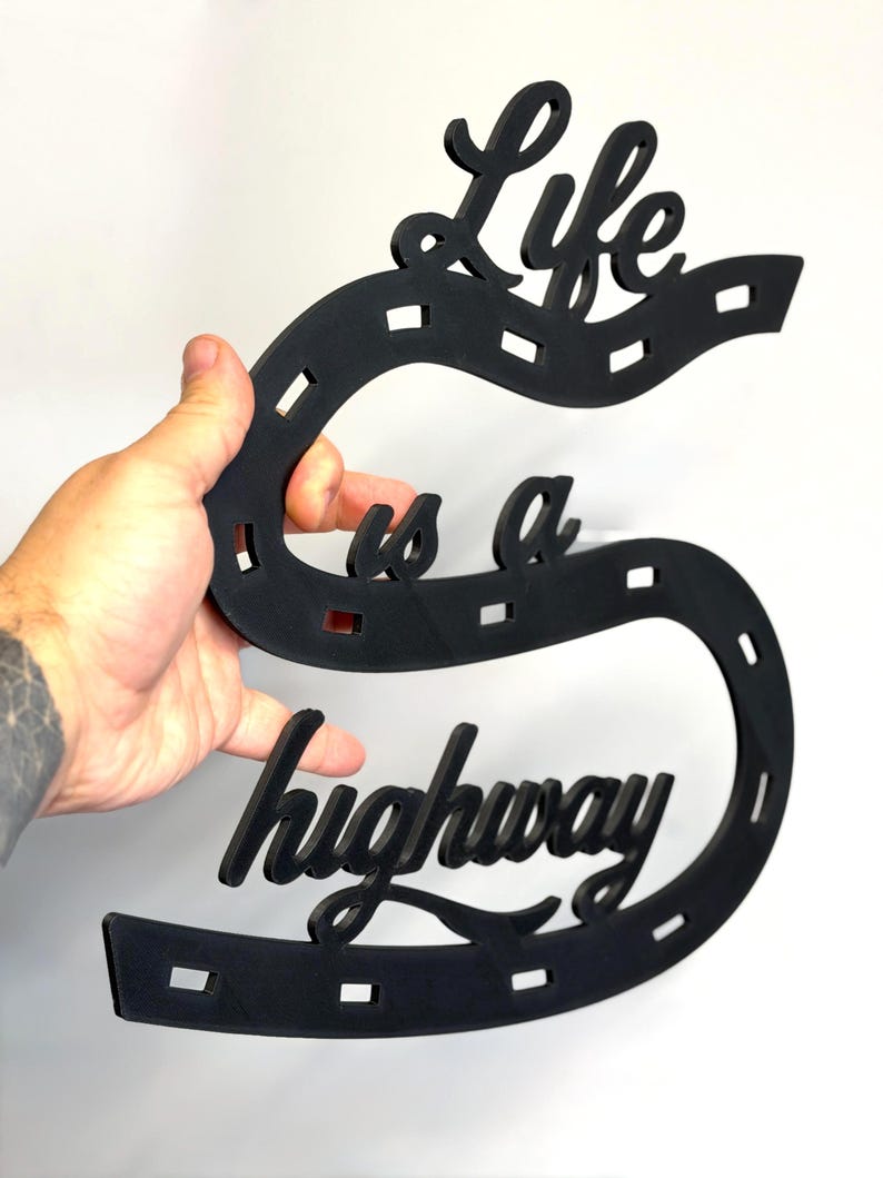 May include: A black wall sign shaped like a winding road with dashed white-style markings, featuring the cut-out text Life is a highway in a cursive script font. The object is constructed from a flat, solid material with a matte finish.