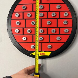 May include: A red and black advent calendar with 24 numbered compartments. The calendar is circular, with a tire-like outer rim. A yellow measuring tape shows the calendar is approximately 18 inches tall.