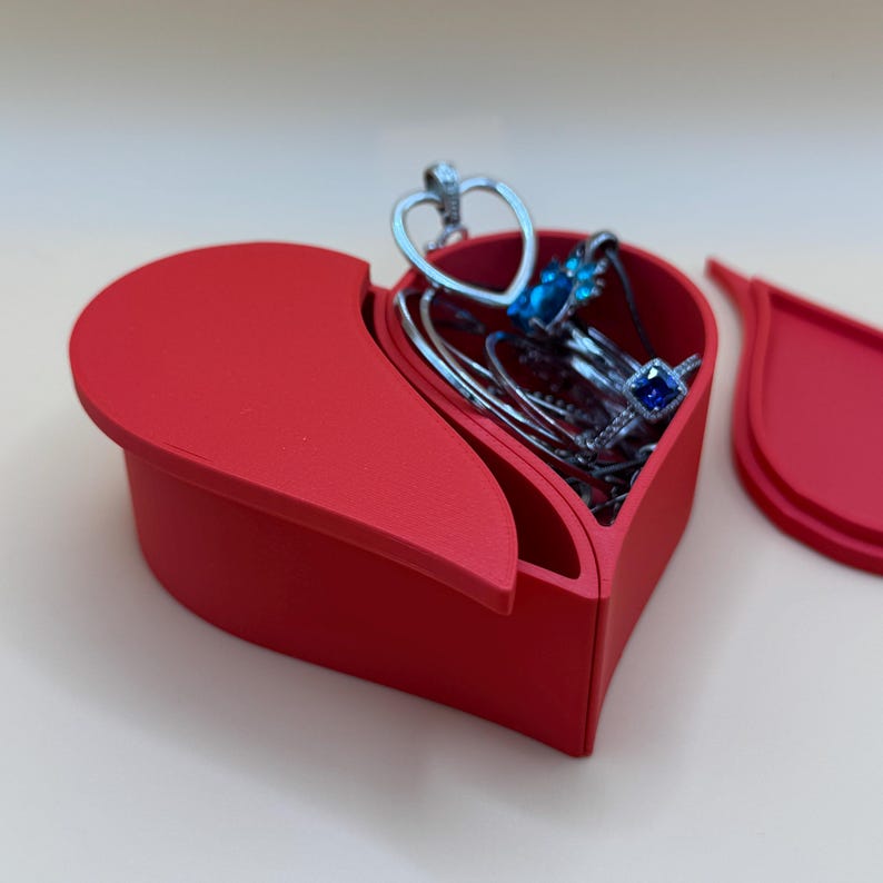 A red heart-shaped box, open to reveal jewellery including bracelets, rings, and a heart-shaped charm. Perfect for Valentine’s Day gifts, keepsakes, or jewellery storage, this split design is made from eco-friendly PLA with a modern, elegant finish.