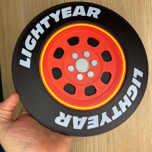 May include: A black and red decorative wheel with the word "LIGHTYEAR" in white lettering. The wheel has a red center with silver accents and a yellow stripe. The item is held by a hand.