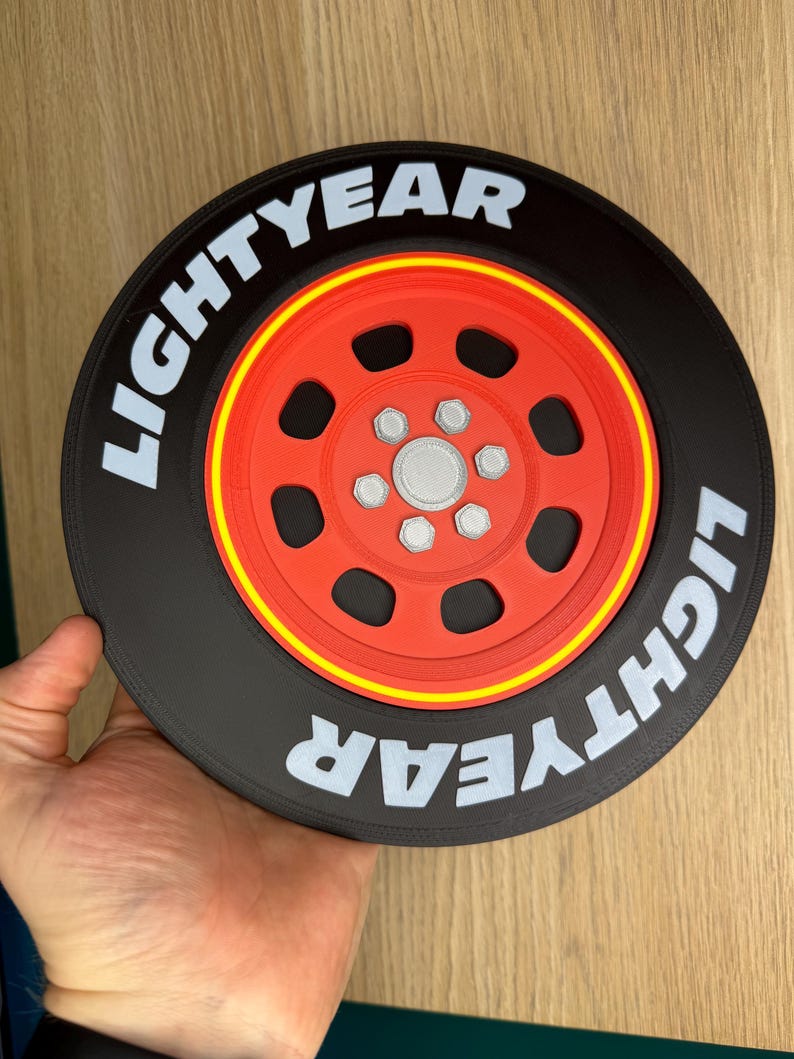 May include: A black and red decorative wheel with the word "LIGHTYEAR" in white lettering. The wheel has a red center with silver accents and a yellow stripe. The item is held by a hand.