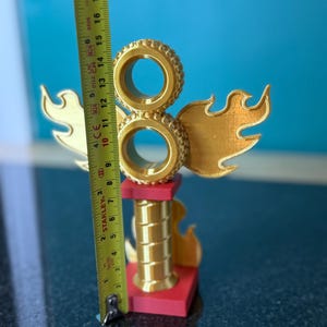 May include: A gold-coloured trophy with a flame design and two stacked tyre shapes. The trophy is approximately 15 cm tall and has a red base. The background is a solid blue.