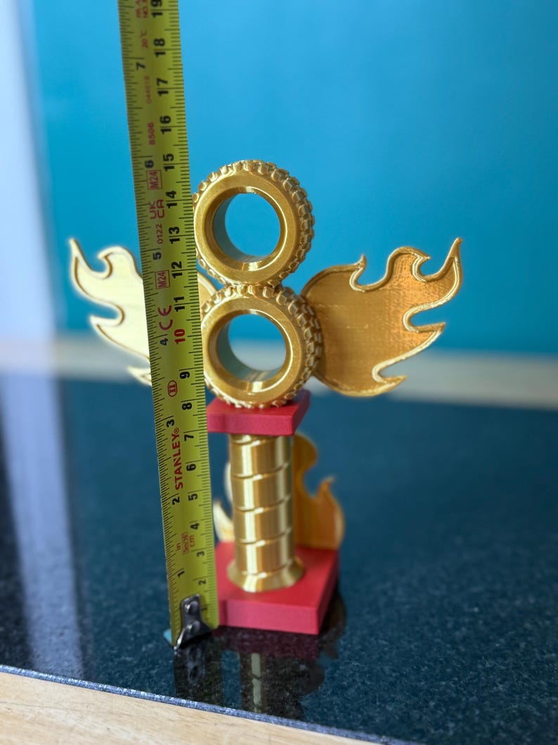 May include: A gold-coloured trophy with a flame design and two stacked tyre shapes. The trophy is approximately 15 cm tall and has a red base. The background is a solid blue.
