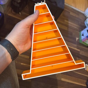 Op de afbeelding: Een feloranje plastic speelgoedbaan met witte details, vastgehouden in een hand. De baan heeft een kegelvorm met verschillende horizontale witte lijnen die niveaus cre&euml;ren. De baan is ontworpen voor speelgoedauto's, met afmetingen in cm.