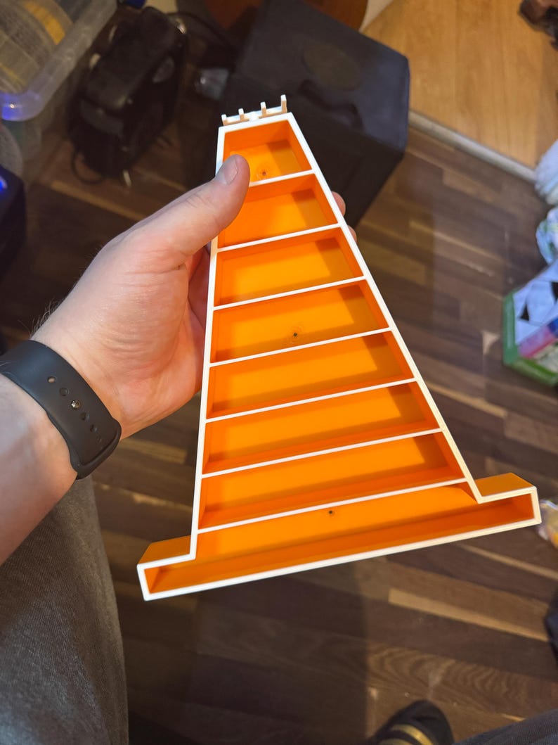 Op de afbeelding: Een feloranje plastic speelgoedbaan met witte details, vastgehouden in een hand. De baan heeft een kegelvorm met verschillende horizontale witte lijnen die niveaus cre&euml;ren. De baan is ontworpen voor speelgoedauto's, met afmetingen in cm.