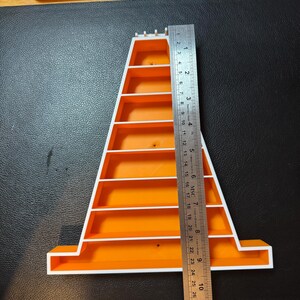 Op de afbeelding: Een oranje en witte plastic plank, ongeveer 28 cm hoog, met meerdere horizontale planken. Een metalen liniaal is naast de plank geplaatst voor de meting. De plank staat op een zwart oppervlak.