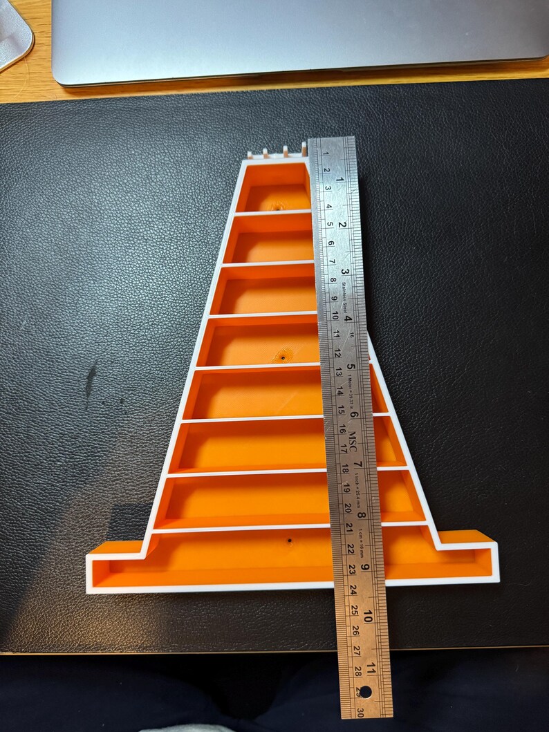 Op de afbeelding: Een oranje en witte plastic plank, ongeveer 28 cm hoog, met meerdere horizontale planken. Een metalen liniaal is naast de plank geplaatst voor de meting. De plank staat op een zwart oppervlak.