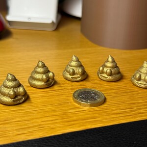 Five gold glittered 2cm tall 3D-printed poop emojis displayed on a wooden surface with a pound coin for scale. Each has a swirled shape, raised eyes, and a smiling mouth. The glitter catches the light for a sparkly effect.