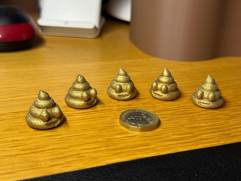 Five gold glittered 2cm tall 3D-printed poop emojis displayed on a wooden surface with a pound coin for scale. Each has a swirled shape, raised eyes, and a smiling mouth. The glitter catches the light for a sparkly effect.