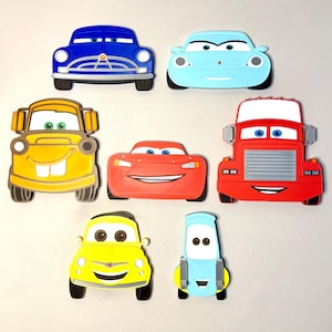 May include: A collection of seven flat, cartoon-style car and truck character ornaments. The set includes a dark blue sedan, a light blue coupe, a yellow tow truck, a red sports car, a red semi-truck, a yellow compact car, and a light blue vehicle.