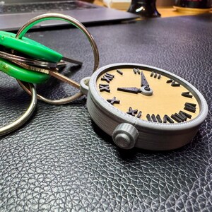 May include: A grey and brown 3D printed clock keyring with Roman numeral markings on the face. The clock is attached to a silver key ring with a green key fob.
