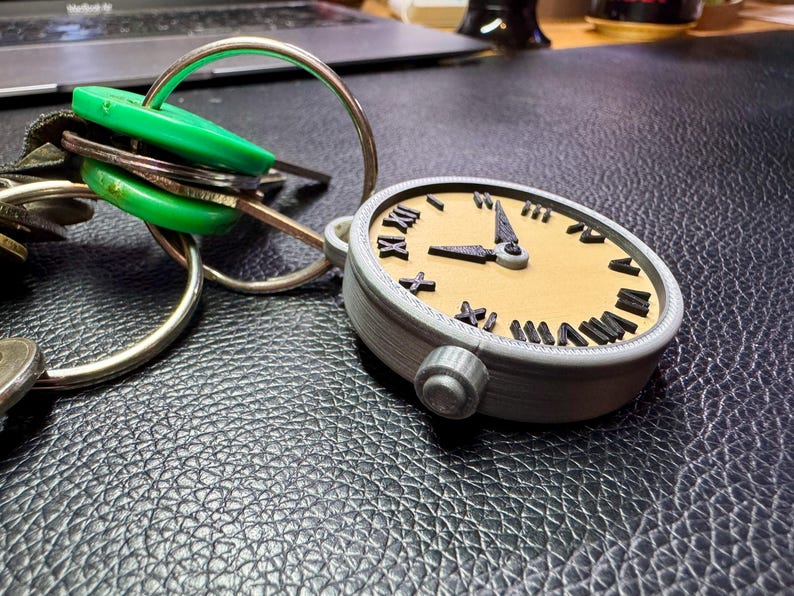 May include: A grey and brown 3D printed clock keyring with Roman numeral markings on the face. The clock is attached to a silver key ring with a green key fob.