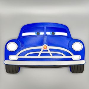 Pode incluir: Um item decorativo azul em forma de carro com detalhes brancos, far&oacute;is e um detalhe central vermelho. O carro tem olhos de desenho animado e uma superf&iacute;cie texturizada, lembrando um ve&iacute;culo vintage. Os pneus s&atilde;o pretos.