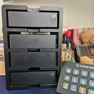 May include: A black 3D-printed vertical storage rack holding four rectangular external hard drives, with the Seagate logo visible on the top and bottom units. Next to the rack is a black Stream Deck controller with backlit keys.