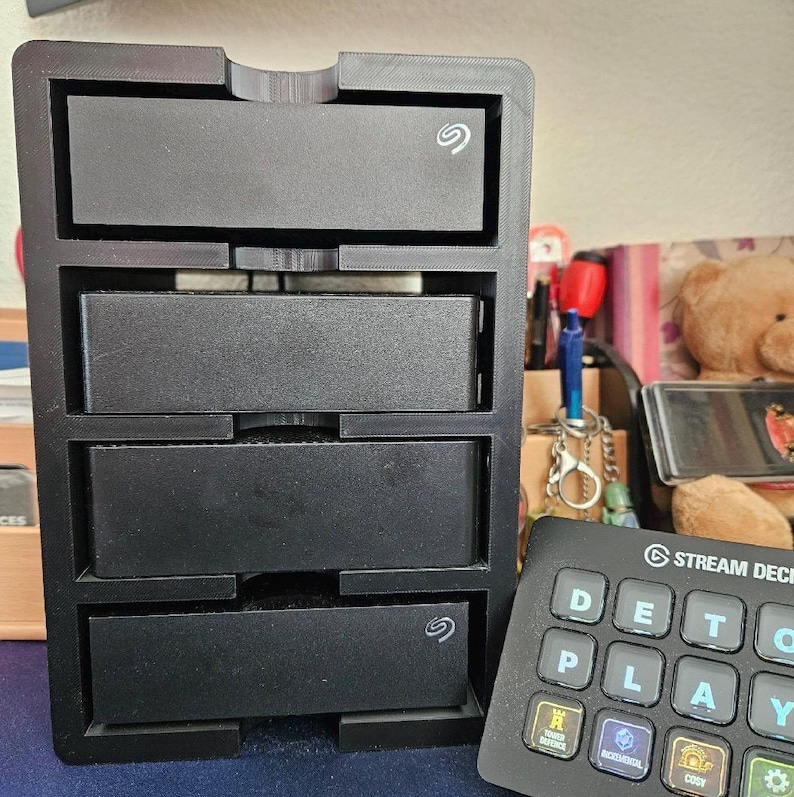 May include: A black 3D-printed vertical storage rack holding four rectangular external hard drives, with the Seagate logo visible on the top and bottom units. Next to the rack is a black Stream Deck controller with backlit keys.