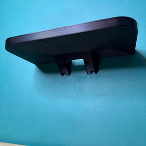May include: A black wall-mounted shelf with a textured surface. The shelf has a rectangular shape with rounded edges and two support brackets. It is mounted on a teal wall.