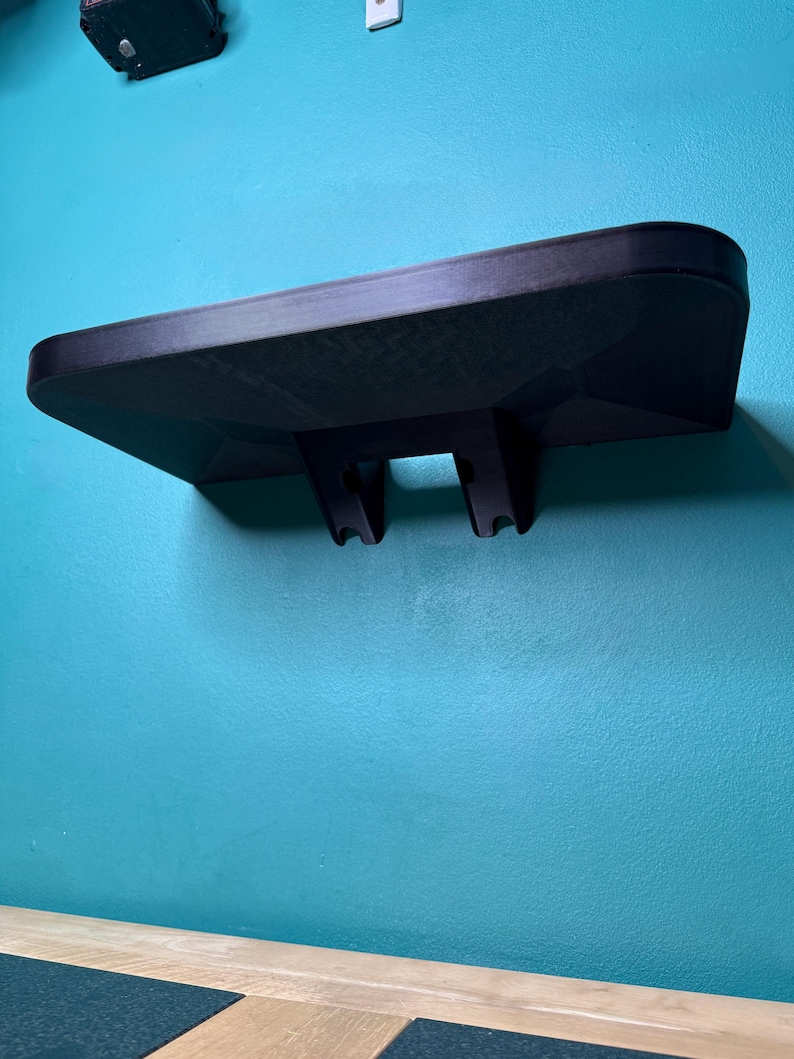 May include: A black wall-mounted shelf with a textured surface. The shelf has a rectangular shape with rounded edges and two support brackets. It is mounted on a teal wall.