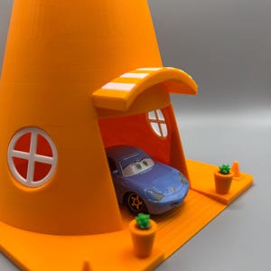 Op de afbeelding: Een blauwe speelgoedauto staat geparkeerd in een feloranje verkeerskegelhuis met een wit gestreept luifel. Het huis heeft twee witte ramen en een klein oranje pad met twee groene planten in potten.