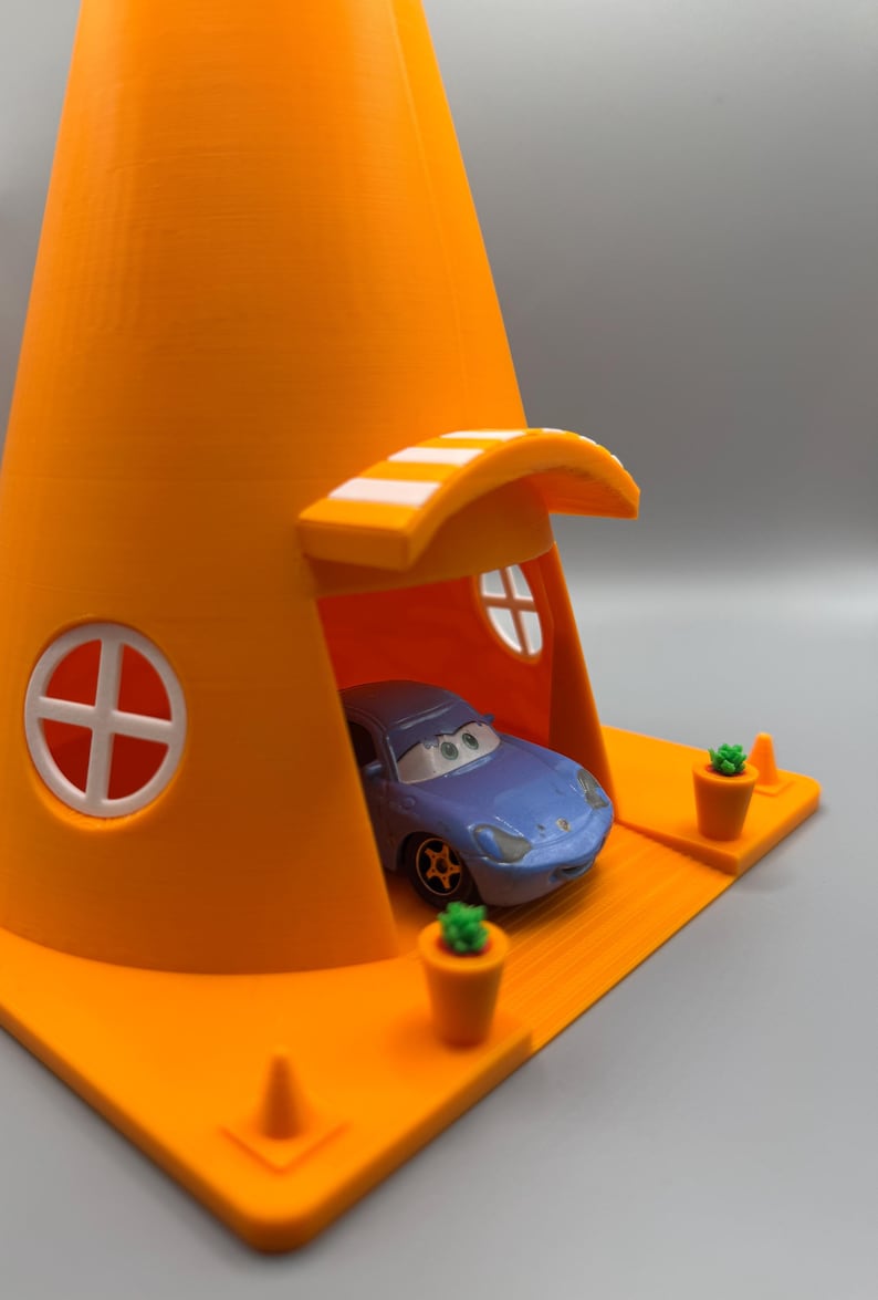 Op de afbeelding: Een blauwe speelgoedauto staat geparkeerd in een feloranje verkeerskegelhuis met een wit gestreept luifel. Het huis heeft twee witte ramen en een klein oranje pad met twee groene planten in potten.