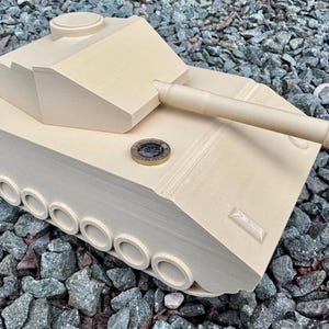 May include: A beige, 3D-printed toy tank model featuring a turret, a long barrel, and a series of circular wheels along the side. A circular coin is placed on the top surface of the tank to provide a sense of scale for the object.