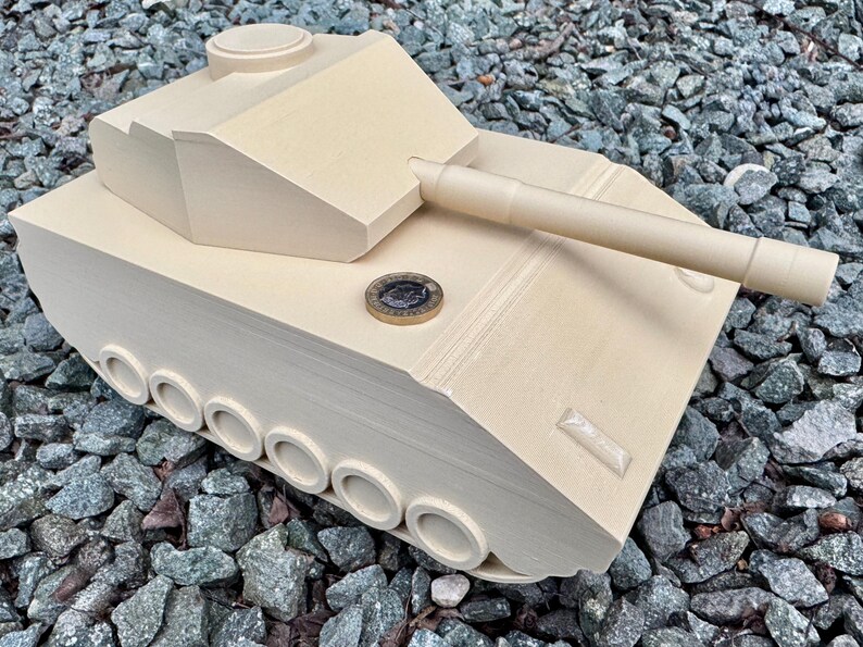 May include: A beige, 3D-printed toy tank model featuring a turret, a long barrel, and a series of circular wheels along the side. A circular coin is placed on the top surface of the tank to provide a sense of scale for the object.