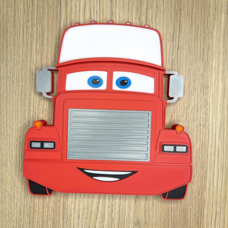 May include: A red cartoon truck with a white windshield, blue eyes, and a smiling mouth. The truck has a silver grill and side mirrors. The design is a flat, two-dimensional style, likely a decorative item or toy.