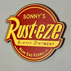 Puede incluir: Un letrero redondo de madera con fondo rojo y letras amarillas. El letrero dice "SONNY'S Rust-eze Bumper Ointment Rear End Formula" en una fuente estilizada. El letrero tiene un borde negro y est&aacute; montado en una pared gris.