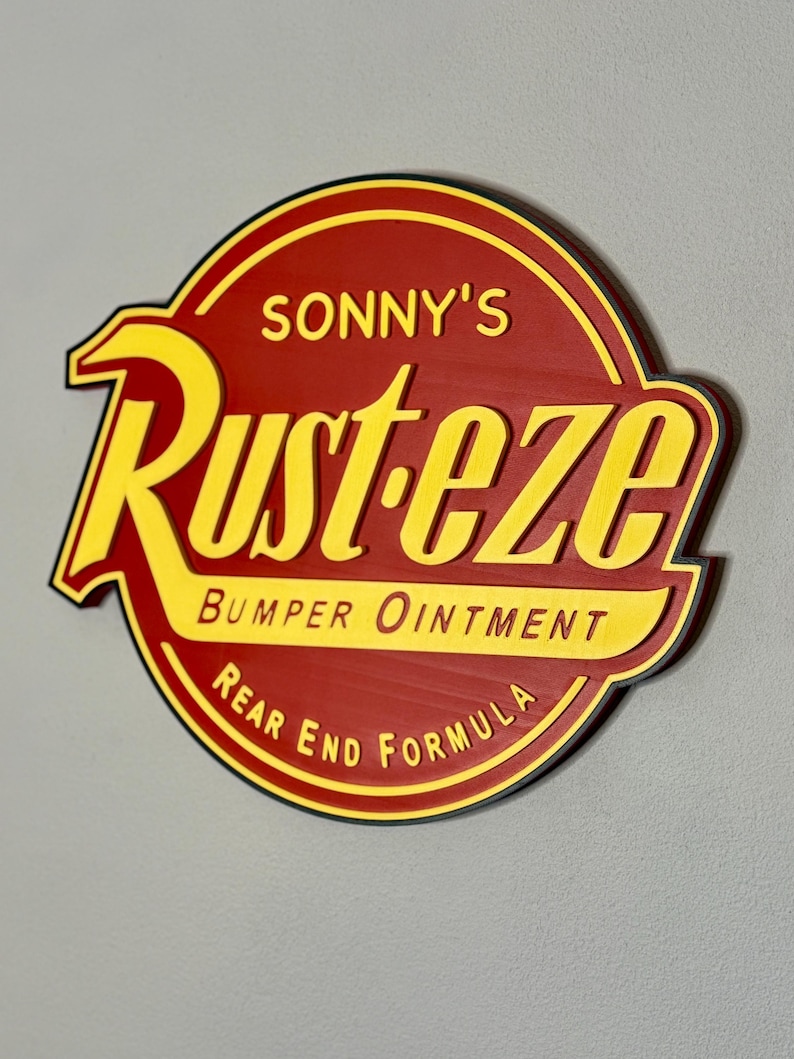 Puede incluir: Un letrero redondo de madera con fondo rojo y letras amarillas. El letrero dice "SONNY'S Rust-eze Bumper Ointment Rear End Formula" en una fuente estilizada. El letrero tiene un borde negro y est&aacute; montado en una pared gris.