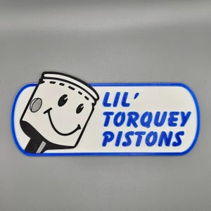 May include: A white and blue patch with the text "LIL' TORQUEY PISTONS". The patch features a smiling piston character in black and white. The patch has a blue border.