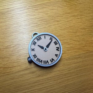 May include: A silver-coloured pocket watch with Roman numeral markings on the face. The watch face is beige with black hands and a black centre.