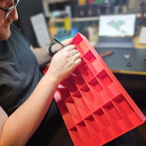 May include: A person working on a bright red, rectangular organizer with multiple compartments. The person is wearing glasses and a headlamp, holding a small tool. The organizer appears to be made of plastic and is being worked on in a workshop setting.