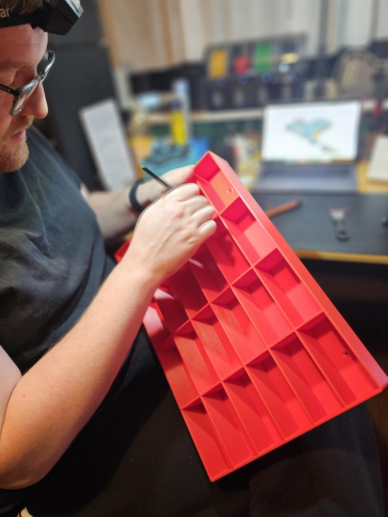 May include: A person working on a bright red, rectangular organizer with multiple compartments. The person is wearing glasses and a headlamp, holding a small tool. The organizer appears to be made of plastic and is being worked on in a workshop setting.