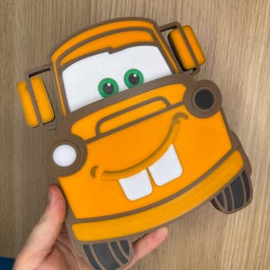 May include: A yellow, orange, and brown plastic truck-shaped object with a wide, smiling face, large white eyes with green pupils, and two prominent white front teeth. The design features dark brown trim, black tires, and orange side mirrors.