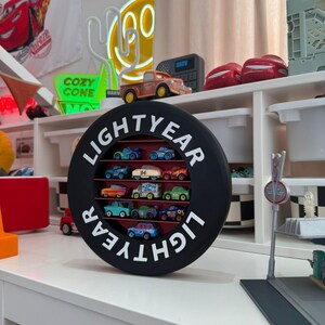 May include: A black circular display case with the word "LIGHTYEAR" in white lettering, holding a collection of miniature toy cars. The cars are various colors and designs, arranged in rows within the case. The display sits on a white surface.