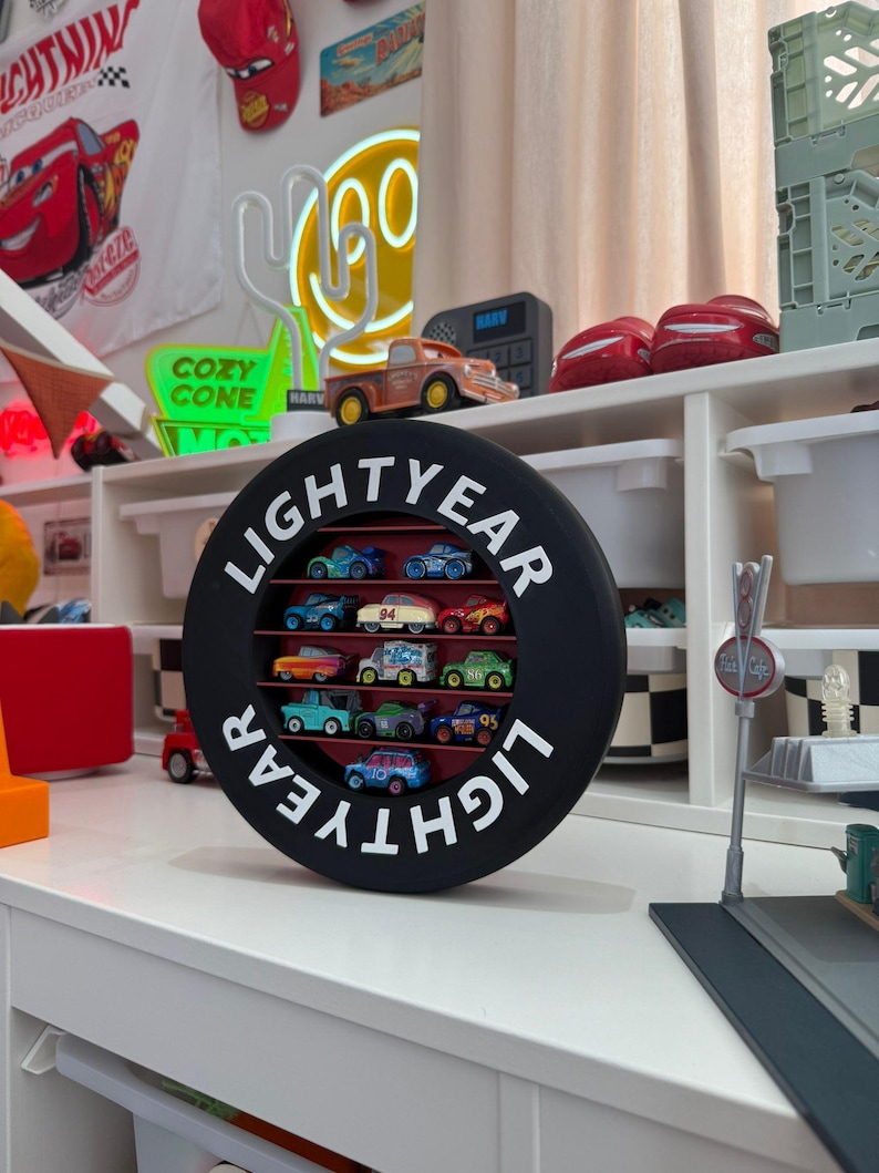 May include: A black circular display case with the word "LIGHTYEAR" in white lettering, holding a collection of miniature toy cars. The cars are various colors and designs, arranged in rows within the case. The display sits on a white surface.