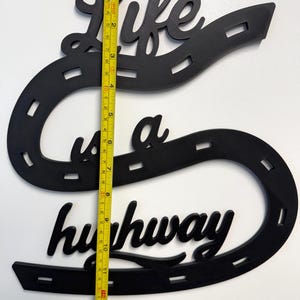 May include: A black wall sign featuring the text Life is a highway shaped like a winding road with dashed lane markings. A yellow measuring tape is placed vertically over the sign, showing a total height of approximately 13.5 inches.