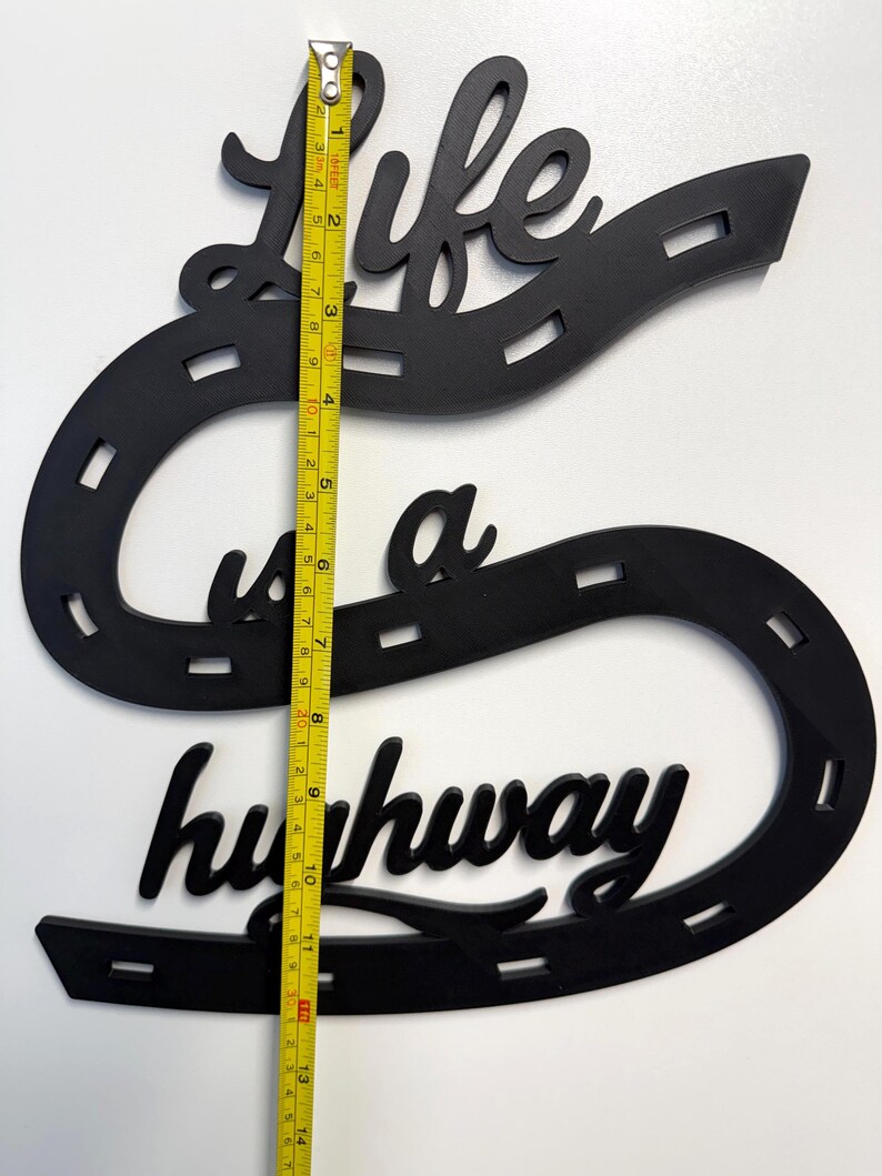 May include: A black wall sign featuring the text Life is a highway shaped like a winding road with dashed lane markings. A yellow measuring tape is placed vertically over the sign, showing a total height of approximately 13.5 inches.