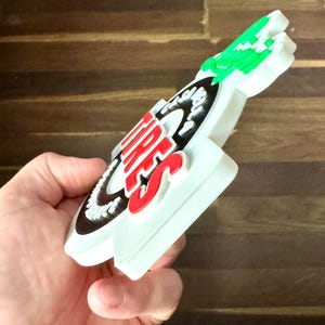 May include: A white, red, black, and green tire-shaped sign with the word "TIRES" in red. The sign is held in a hand and has a unique shape with a green accent at the top.