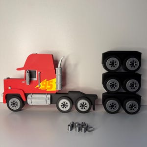 May include: A red toy truck with the number 95 and a yellow lightning bolt graphic on the side. The truck has a black chassis and silver wheels. There are three sets of black wheels and silver cylinders.